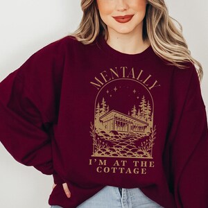 May include: Burgundy sweatshirt with a gold graphic. The graphic features a cottage scene with the text "MENTALLY I'M AT THE COTTAGE". The sweatshirt is a crew neck style and has long sleeves. The overall theme is cozy and relaxed.