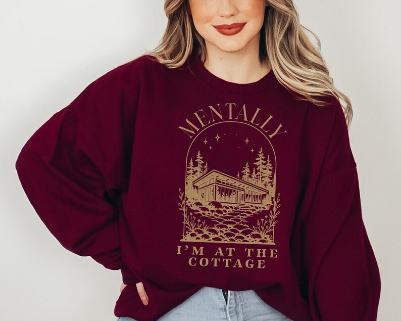 May include: Burgundy sweatshirt with a gold graphic. The graphic features a cottage scene with the text "MENTALLY I'M AT THE COTTAGE". The sweatshirt is a crew neck style and has long sleeves. The overall theme is cozy and relaxed.