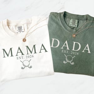 May include: Two t-shirts, one cream and one olive green, with the words "MAMA" and "DADA" printed on them. Each shirt has "EST. 2026" and a golf ball and club graphic. Gold necklaces with round pendants are draped on the shirts.