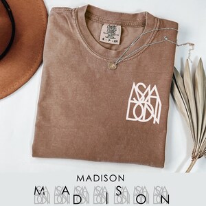 May include: A brown Comfort Colors t-shirt with the name "MADISON" printed in white. The shirt has a crew neck and short sleeves. A silver necklace with a small pendant is draped across the shirt. A brown hat and dried plant are in the background.
