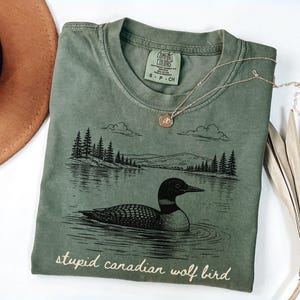 May include: Sage green t-shirt with a graphic of a loon on a lake, trees, and mountains. The text "stupid canadian wolf bird" is printed below the graphic. A gold necklace and a brown hat are also in the image.