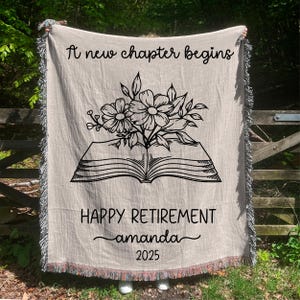 May include: Beige woven throw blanket with black text and floral design. The blanket reads "A new chapter begins" at the top, and "HAPPY RETIREMENT amanda 2025" at the bottom. The design includes an open book with flowers. The edges have a fringed trim.
