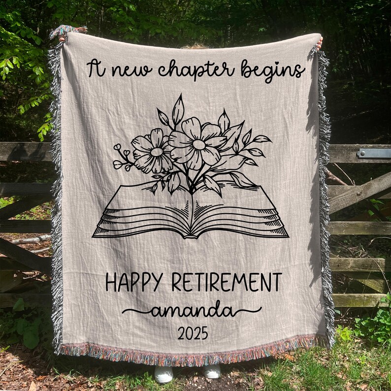 May include: Beige woven throw blanket with black text and floral design. The blanket reads "A new chapter begins" at the top, and "HAPPY RETIREMENT amanda 2025" at the bottom. The design includes an open book with flowers. The edges have a fringed trim.