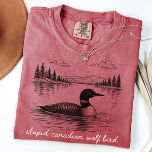 May include: A red-pink t-shirt with a graphic of a loon on a lake, with trees and mountains in the background. The text "stupid canadian wolf bird" is printed at the bottom. A silver necklace with a pendant is on the shirt.