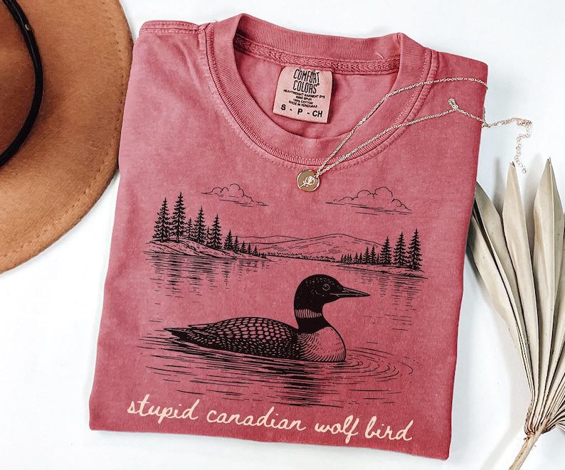 May include: A red-pink t-shirt with a graphic of a loon on a lake, with trees and mountains in the background. The text "stupid canadian wolf bird" is printed at the bottom. A silver necklace with a pendant is on the shirt.