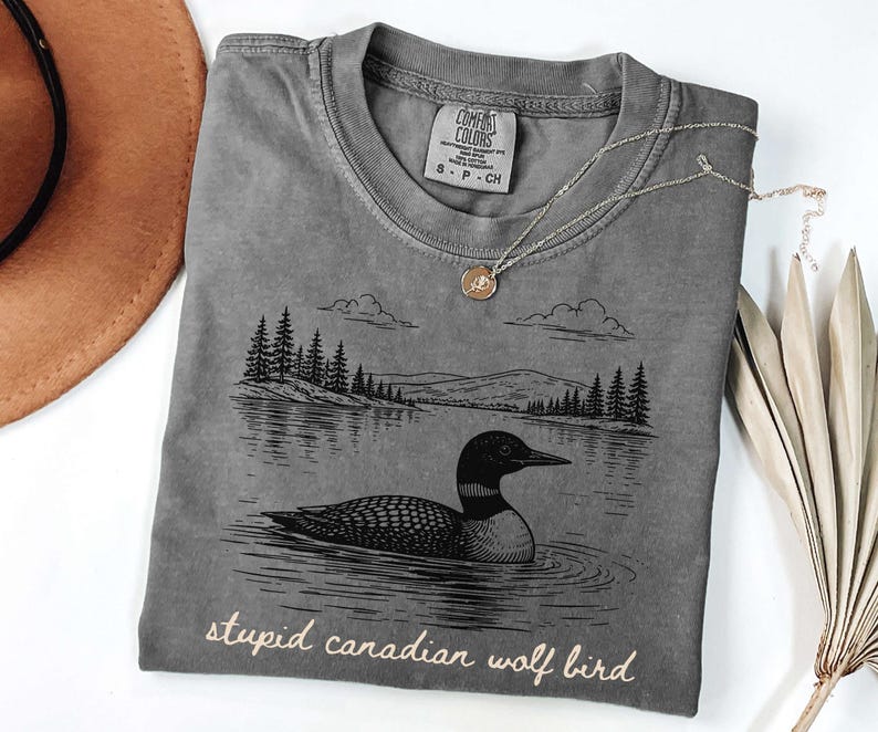 May include: Gray t-shirt with a graphic of a loon on a lake, trees, and mountains. The text "stupid canadian wolf bird" is printed at the bottom. A gold necklace and a brown hat are also in the image.