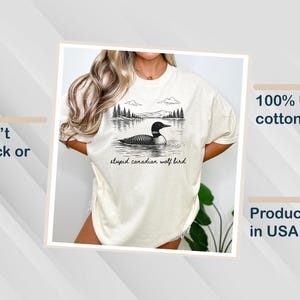 May include: Cream-colored t-shirt featuring a black and white illustration of a loon on a lake with trees and mountains in the background. The text "stupid canadian wolf bird" is below the image. Made of 100% US cotton.