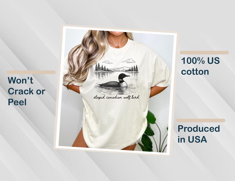 May include: Cream-colored t-shirt featuring a black and white illustration of a loon on a lake with trees and mountains in the background. The text "stupid canadian wolf bird" is below the image. Made of 100% US cotton.