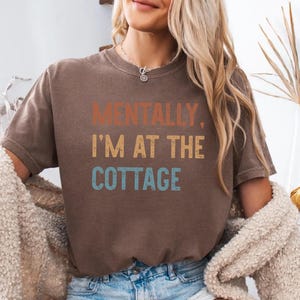 May include: A brown t-shirt with the text "MENTALLY, I'M AT THE COTTAGE" in orange, yellow, and blue. The shirt is a casual style, suitable for everyday wear.