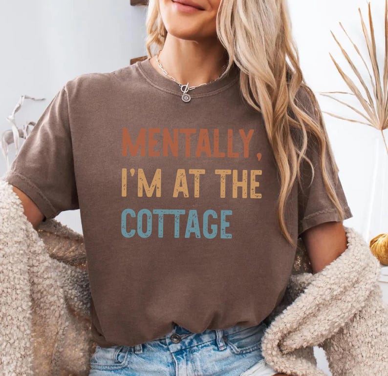 May include: A brown t-shirt with the text "MENTALLY, I'M AT THE COTTAGE" in orange, yellow, and blue. The shirt is a casual style, suitable for everyday wear.