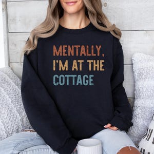 May include: Black sweatshirt with the text "MENTALLY, I'M AT THE COTTAGE" in orange, yellow, and teal lettering. The sweatshirt is worn by a person with blonde hair, sitting on a couch with white pillows.