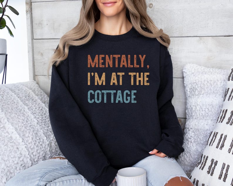 May include: Black sweatshirt with the text "MENTALLY, I'M AT THE COTTAGE" in orange, yellow, and teal lettering. The sweatshirt is worn by a person with blonde hair, sitting on a couch with white pillows.