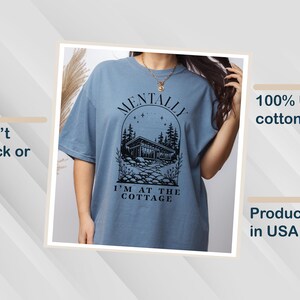 May include: A blue t-shirt with the text "MENTALLY I'M AT THE COTTAGE" in black. The graphic features a cabin scene with trees and stars. The shirt is made of 100% US cotton and is produced in the USA. The shirt is designed to not crack or peel.