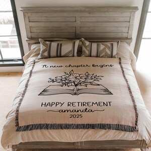May include: A beige throw blanket with a black floral and book design, and the text "A new chapter begins" and "HAPPY RETIREMENT amanda 2025". The blanket has a fringed edge and is displayed on a bed with pillows.