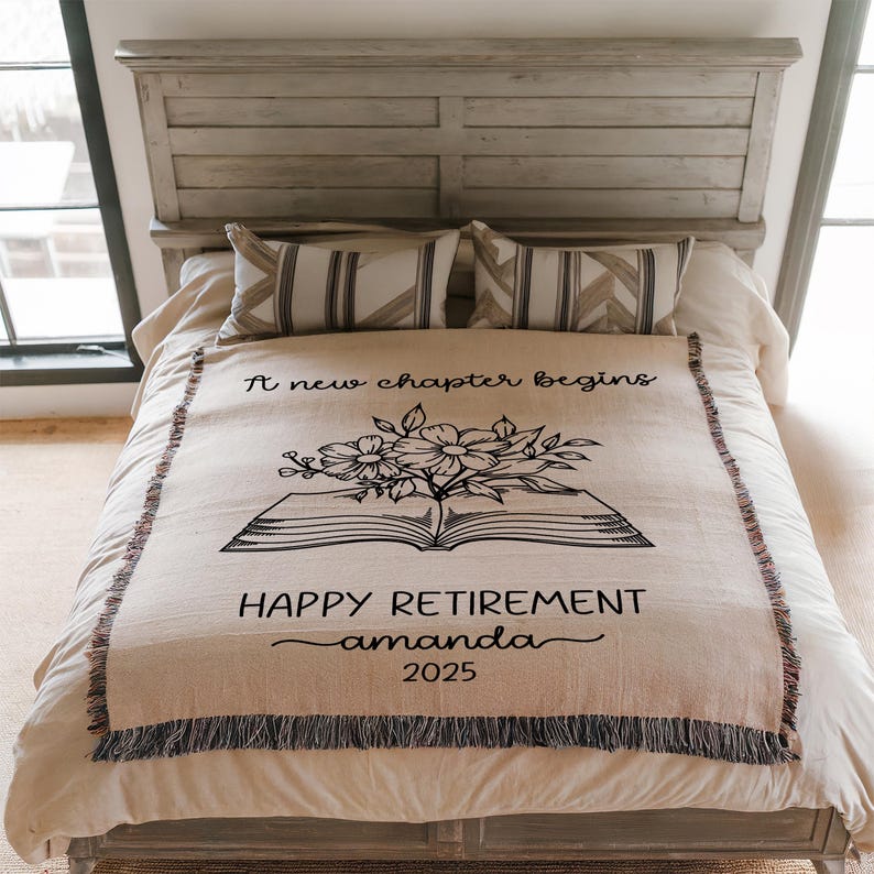 May include: A beige throw blanket with a black floral and book design, and the text "A new chapter begins" and "HAPPY RETIREMENT amanda 2025". The blanket has a fringed edge and is displayed on a bed with pillows.