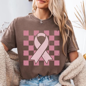 May include: Brown t-shirt with a pink ribbon design on a pink and brown chequered background. The ribbon is a symbol of breast cancer awareness. The shirt is a casual style.