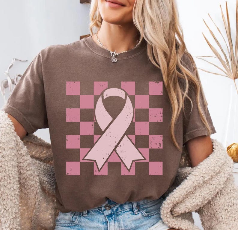 May include: Brown t-shirt with a pink ribbon design on a pink and brown chequered background. The ribbon is a symbol of breast cancer awareness. The shirt is a casual style.