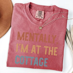 May include: A red Comfort Colors t-shirt with the text "MENTALLY, I'M AT THE COTTAGE" in brown, orange, and blue. A gold necklace and a brown hat are also visible.