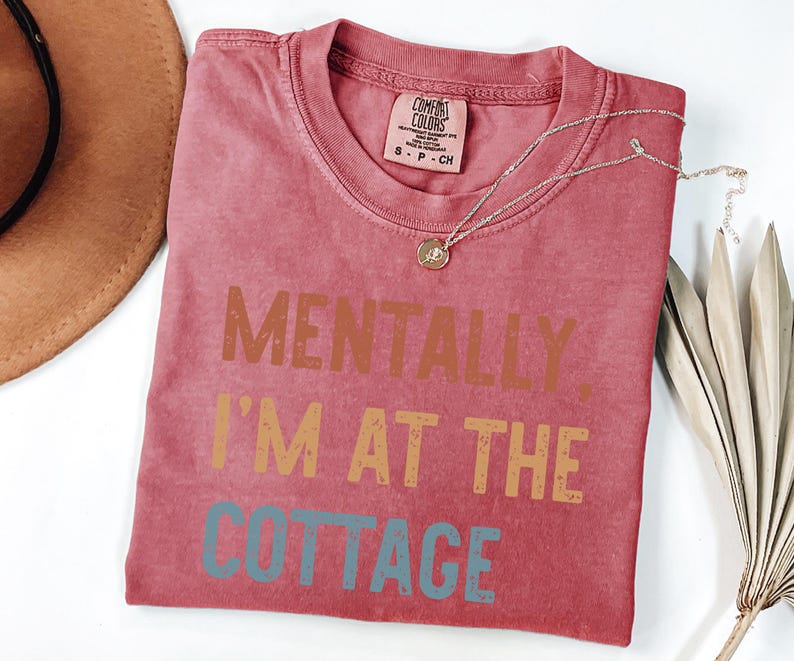 May include: A red Comfort Colors t-shirt with the text "MENTALLY, I'M AT THE COTTAGE" in brown, orange, and blue. A gold necklace and a brown hat are also visible.