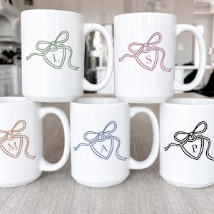 May include: Six white ceramic mugs with a large handle are arranged on a light-colored surface. Each mug features a different colored bow and ribbon design with a letter inside a shield. The letters are L, S, M, A, and P.