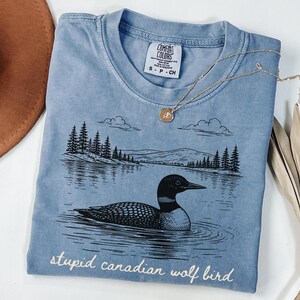May include: A light blue Comfort Colors t-shirt with a graphic of a loon swimming in a lake, with trees and mountains in the background. The text "stupid canadian wolf bird" is printed at the bottom. A gold necklace is draped across the shirt.