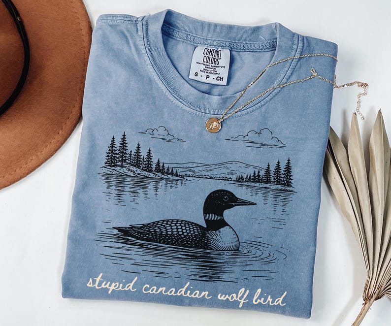 May include: A light blue Comfort Colors t-shirt with a graphic of a loon swimming in a lake, with trees and mountains in the background. The text "stupid canadian wolf bird" is printed at the bottom. A gold necklace is draped across the shirt.