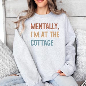May include: A light gray sweatshirt with the text "MENTALLY, I'M AT THE COTTAGE" in brown, tan, and teal lettering. The sweatshirt is a long-sleeved crew neck.