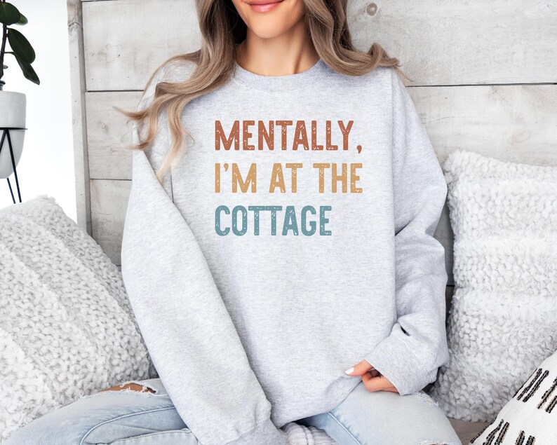May include: A light gray sweatshirt with the text "MENTALLY, I'M AT THE COTTAGE" in brown, tan, and teal lettering. The sweatshirt is a long-sleeved crew neck.