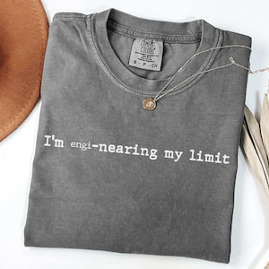 May include: A gray Comfort Colors t-shirt with the text "I'm engi-nearing my limit" in white. A gold necklace with a pendant is draped across the shirt. A brown hat and dried plant are in the background.