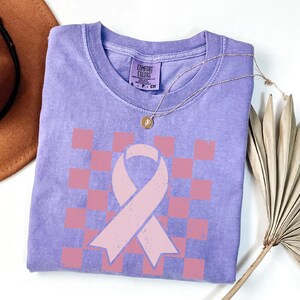 May include: A lilac Comfort Colors t-shirt featuring a pink ribbon and chequered pattern. The shirt has a crew neck and short sleeves. A gold necklace with a pendant is draped across the shirt. A brown hat and dried palm frond are in the background.