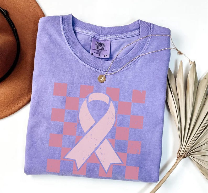 May include: A lilac Comfort Colors t-shirt featuring a pink ribbon and chequered pattern. The shirt has a crew neck and short sleeves. A gold necklace with a pendant is draped across the shirt. A brown hat and dried palm frond are in the background.