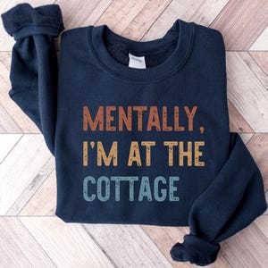 May include: Navy blue sweatshirt with the text "MENTALLY, I'M AT THE COTTAGE" in orange, yellow, and blue. The sweatshirt has a crew neck and long sleeves with ribbed cuffs. The garment is displayed on a light wood-grain surface.