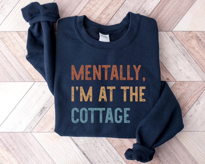 May include: Navy blue sweatshirt with the text "MENTALLY, I'M AT THE COTTAGE" in orange, yellow, and blue. The sweatshirt has a crew neck and long sleeves with ribbed cuffs. The garment is displayed on a light wood-grain surface.