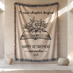 May include: Beige woven throw blanket with a decorative fringe. The blanket features the text "A new chapter begins" and "HAPPY RETIREMENT amanda 2025" with a floral and book illustration in black.
