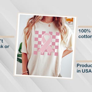 May include: A cream-coloured t-shirt with a pink ribbon design on a checkered pattern. Made from 100% US cotton and produced in the USA. The print is designed to resist cracking or peeling.