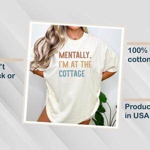 May include: Cream-colored t-shirt with the text "MENTALLY, I'M AT THE COTTAGE" in brown, blue, and orange. The shirt is made of 100% US cotton and is produced in the USA. The design is made to not crack or peel.