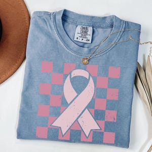 May include: A blue Comfort Colors t-shirt featuring a pink ribbon design on a pink and white chequered pattern. A gold necklace with a pendant is draped over the shirt. A brown hat and dried plant are in the background.