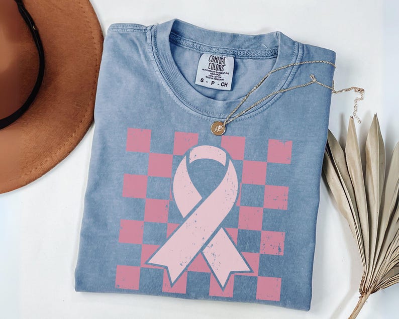 May include: A blue Comfort Colors t-shirt featuring a pink ribbon design on a pink and white chequered pattern. A gold necklace with a pendant is draped over the shirt. A brown hat and dried plant are in the background.