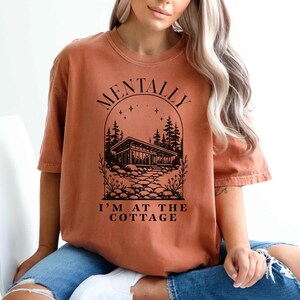 May include: Burnt orange t-shirt with the text "MENTALLY I'M AT THE COTTAGE" in black. The graphic features a cabin scene with trees and a stone path, all within an arched design. The shirt has short sleeves and a relaxed fit.
