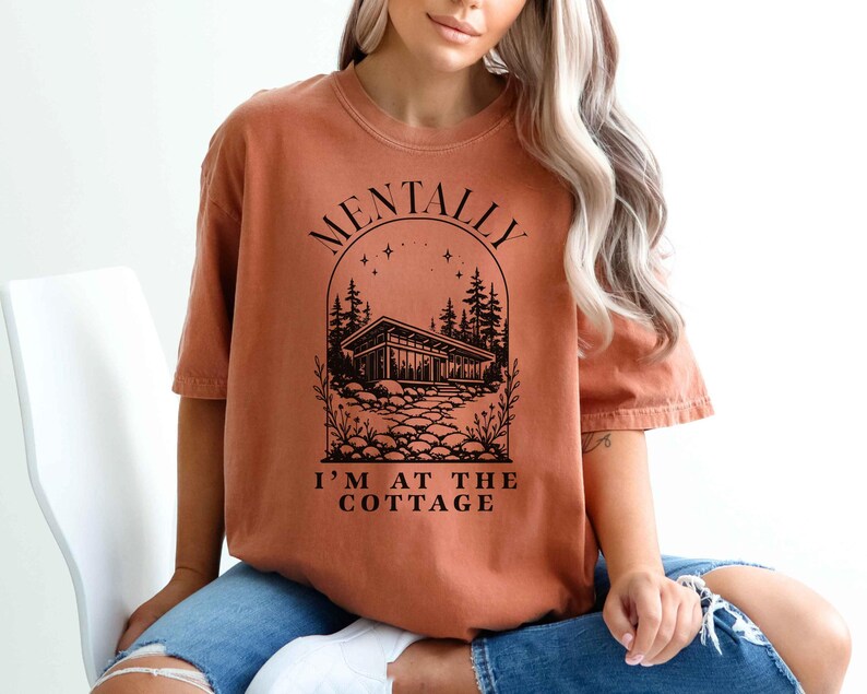 May include: Burnt orange t-shirt with the text "MENTALLY I'M AT THE COTTAGE" in black. The graphic features a cabin scene with trees and a stone path, all within an arched design. The shirt has short sleeves and a relaxed fit.