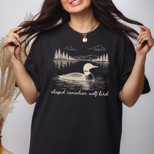 May include: Black t-shirt featuring a graphic of a loon swimming in a lake with trees and mountains in the background. The text "stupid canadian wolf bird" is printed below the graphic.