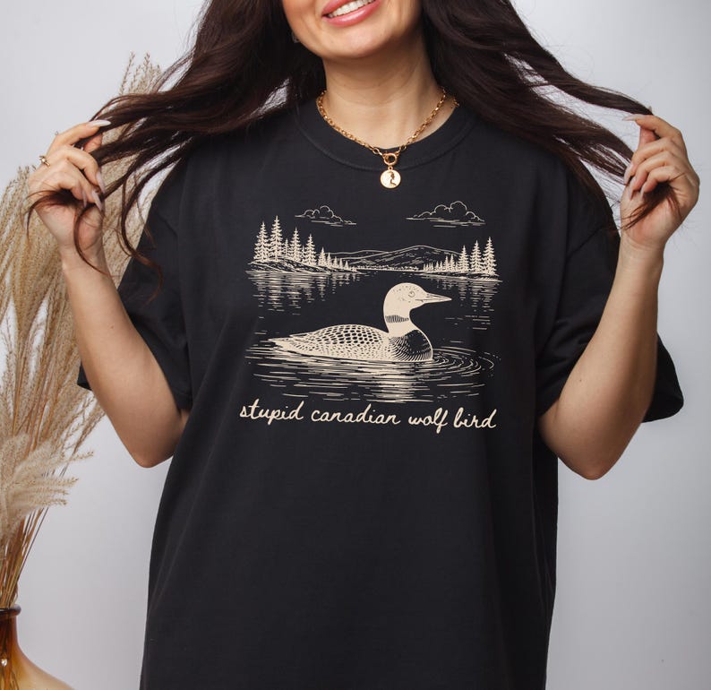 May include: Black t-shirt featuring a graphic of a loon swimming in a lake with trees and mountains in the background. The text "stupid canadian wolf bird" is printed below the graphic.