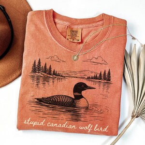 May include: Coral-colored t-shirt with a graphic of a loon on a lake, trees, and mountains. The text "stupid canadian wolf bird" is printed at the bottom. A gold necklace and a brown hat are also in the image.
