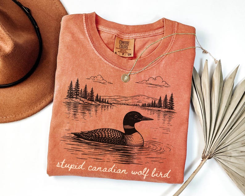 May include: Coral-colored t-shirt with a graphic of a loon on a lake, trees, and mountains. The text "stupid canadian wolf bird" is printed at the bottom. A gold necklace and a brown hat are also in the image.