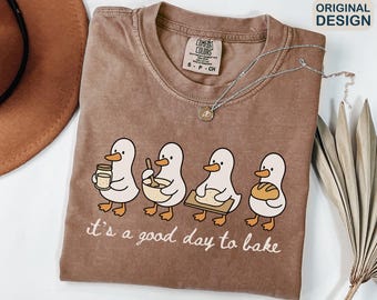 May include: A brown Comfort Colors t-shirt featuring a graphic of four cartoon ducks baking. The ducks are holding a jar, mixing bowl, cutting board, and loaf of bread. The text "it's a good day to bake" is below the ducks. A silver necklace is draped across the shirt.