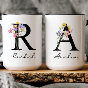 May include: Two white ceramic mugs with black handles and rims. Each mug features a large black letter, "R" and "A", adorned with colorful floral arrangements. The names "Rachel" and "Amelia" are written below the letters.