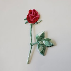 May include: A red rose with green leaves embroidered on a white background. The rose is a single bloom with a stem and leaves.