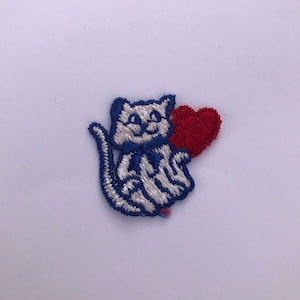 May include: Embroidered patch featuring a white cat with blue accents and a red heart. The cat is sitting and has a blue bow tie. The patch is likely for clothing or accessories.