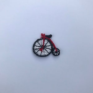 May include: Embroidered patch featuring a vintage bicycle design. The bicycle has a large front wheel and a smaller rear wheel. The wheels are white with black trim, and the frame is red and black. The patch is on a white background.