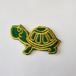 May include: Embroidered turtle patch in green and gold. The turtle has a green body and head with gold embroidered details on the shell and edges. The patch is likely for clothing or accessories.